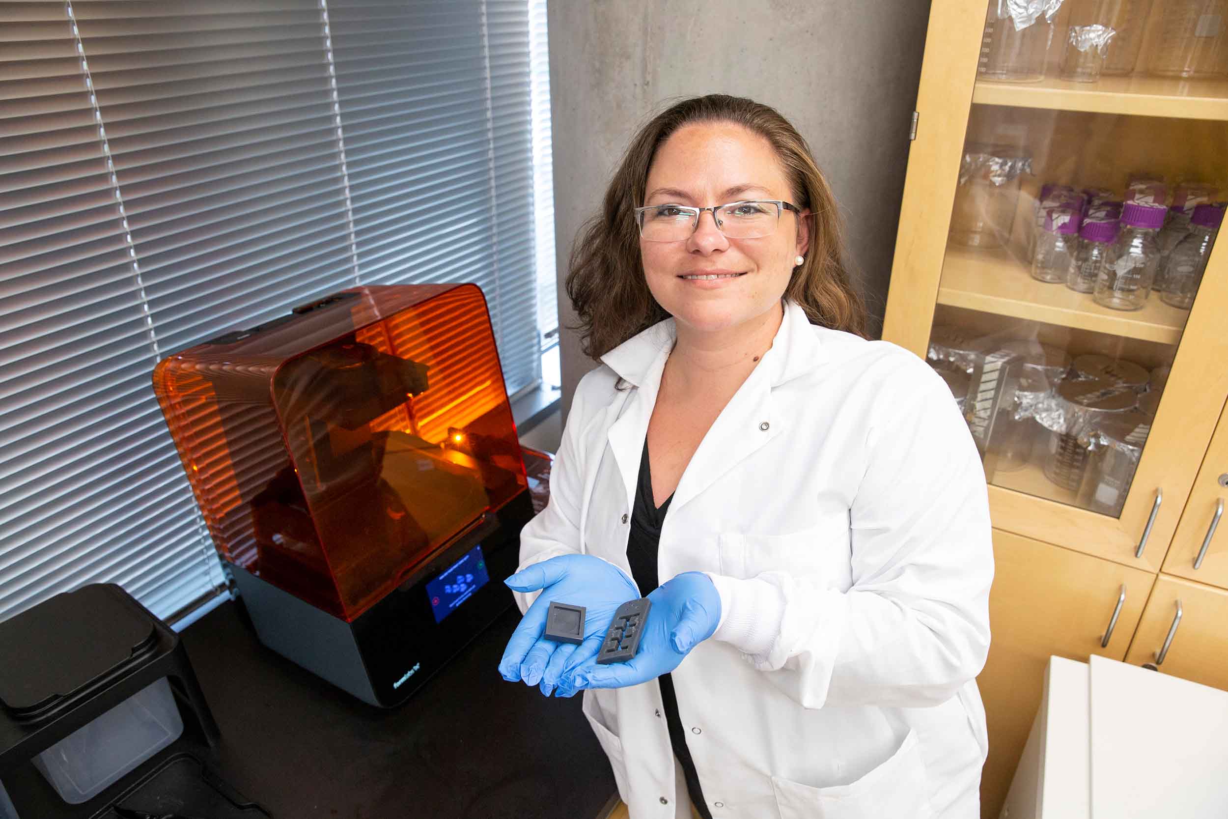 Jessica Weaver, dressed in a lab coat and latex gloves, holding a 3D-printed object