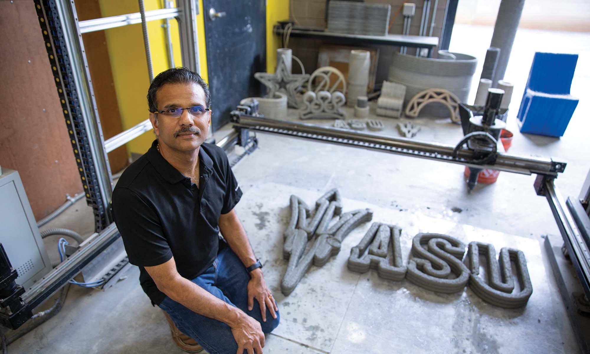 Fulton Professor of Structural Materials Narayanan Neithalath, wearing a black polo shirt and jeans, kneels next to a large, textured 3D print of the letters "ASU" with the pitchfork logo in a workshop.